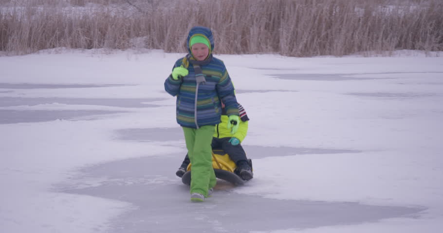 two happy children in warm winter clothes ride on yellow sled on frozen snow-covered river.  Concept of friendly sports family. Winter holidays in village. Christmas eve