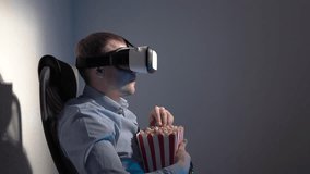 A young man with a box of popcorn in his hand watching a video using a virtual reality headset. Sitting in a chair eating popcorn in a dark room.