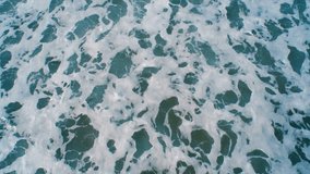 Aerial drone footage of ocean waves crashing on shore. As the waves splash they create a texture from the white sea foam. As the waves calm the textures change. Overhead perspective. HD 1080 video.