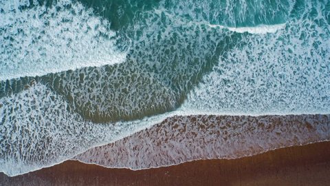 Aerial drone slow motion footage of ocean waves reaching shore. Lockdown shot of sea waves creating a texture from the white sea foam. Footage is filmed from an overhead perspective. HD 1080 video.
