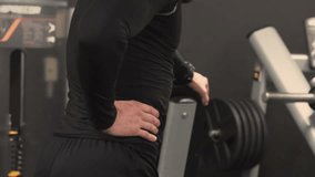 Side view of athletic handsome young man with beard in black sportswear stretching back muscles after exercising in gym, pressing hands against lower back, bending. Health concept
