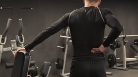 Rear view of athletic handsome young man with beard in black sportswear stretching back muscles after exercising in gym, pressing hands against lower back, bending. Health concept