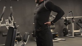 Athletic handsome young man with beard in black sportswear stretching back muscles after exercising in gym, pressing hands against lower back, bending. Health concept