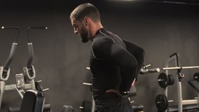 Athletic handsome young man with beard in black sportswear stretching back muscles after exercising in gym, pressing hands against lower back, having backache. Health concept