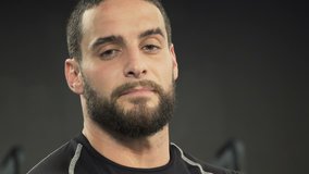 Close up shot of tired athletic handsome young man with beard in black sportswear looking at camera, blinking. Fatigue concept