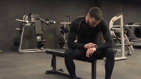 Athletic handsome young man with beard in black sportswear sitting on gym bench after exercising, checking health data on fitness tracker app on smartwatch