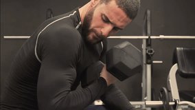 Medium shot of athletic handsome young man with beard in black sportswear flexing arms with dumbbells in his hand, working out in gym. Static shot. Training concept.