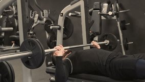 Athletic handsome young man with beard in black sportswear doing bench press with barbell in gym, training hands. Medium shot, static shot, side view. Training concept.
