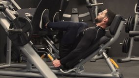 Side view of athletic handsome young man with beard in black sportswear exercising on leg press machine in gym. Long shot, static shot. Training concept.