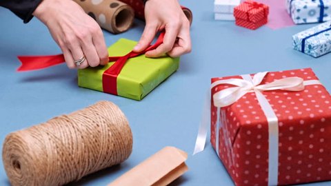Person hands wrapping present on blue background with paper. Woman packaging wrapped gift box with red ribbon and bow. Birthday, anniversary, Valentine's, Christmas holidays packaging.