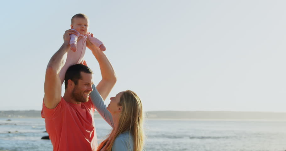 Side view of mid adult Caucasian parents playing with baby at beach on a sunny day. Father carrying baby on shoulder 