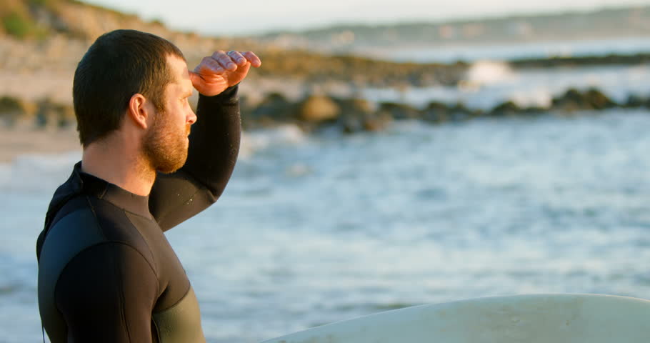 Side view close up of mid-adult Caucasian male surfer with shielding eyes looking at sea at beach. Calm sea in the background 