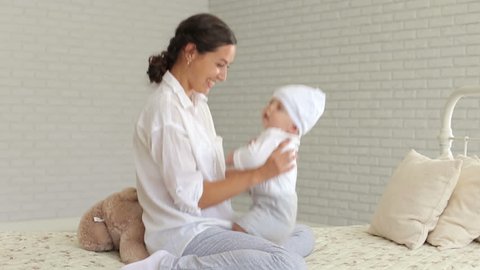 Portrait of a young mother with a newborn baby at home on the bed, they are dressed in white clothes. The concept of a happy motherhood.