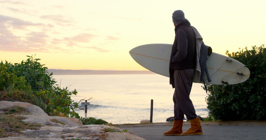Rear view of mid-adult man with surfboard standing on road near seaside. Man looking at sea during sunset 