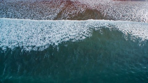 Aerial drone slow motion video of sea waves reaching shore. Lockdown of ocean waves creating a texture from the white sea foam. The footage is filmed from an overhead perspective. HD 1080 video.