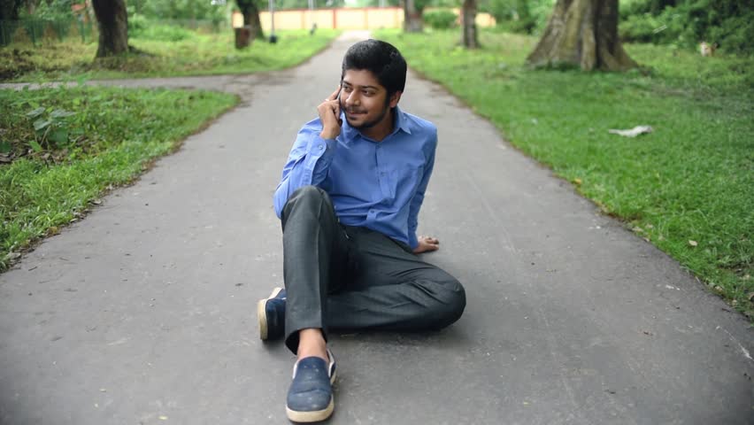 A cheerful and confident Asian man sits on the middle of a road and talks on phone with his beloved and smiles