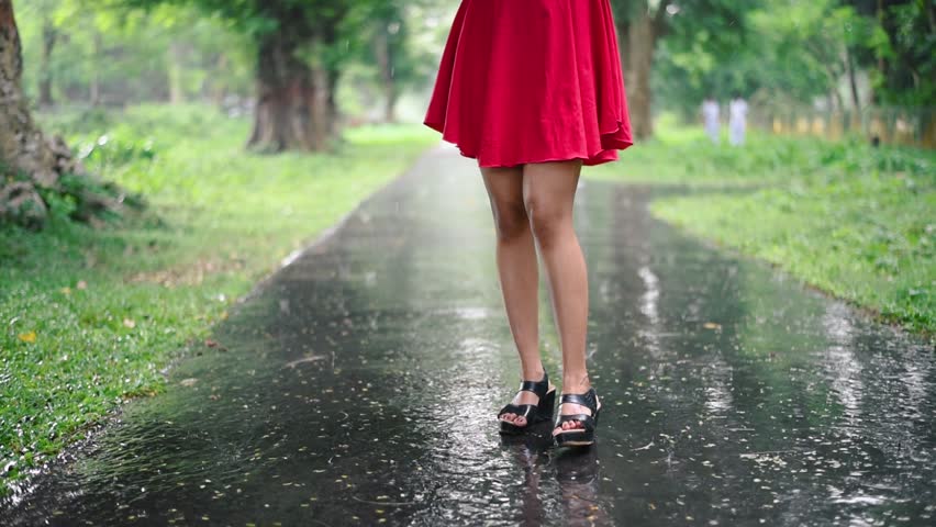 A cute and romantic girl is playing and dancing in Rain on a road outdoor with her umbrella with a short red dress, camera tilt, slow motion