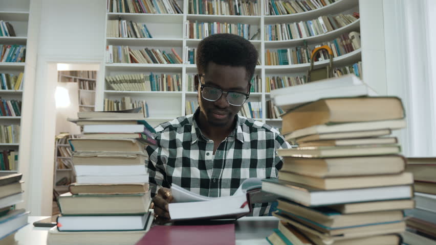 Portrait of african young man in shirt with glasses reading book while sitting in the light modern library. Dolly shot. | Shutterstock HD Video #1023162934