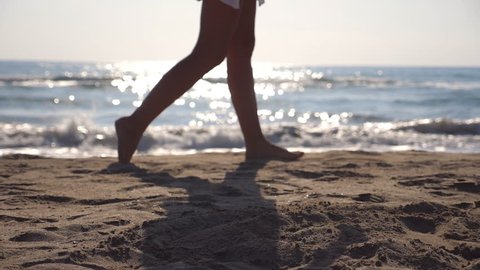Female feet walking at the sea beach on a sunny day with waves at background. Legs of young woman stepping at the sand. Summer vacation or holiday concept. Side view Slow motion Close up