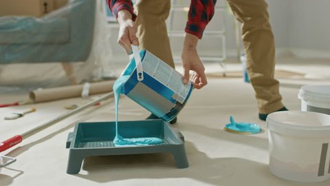 Young Man in Brown Jeans and Red Checked Shirt Picks Up a Can of Light Blue Paint and Pours it Into a Tray. Room Renovations at Home.