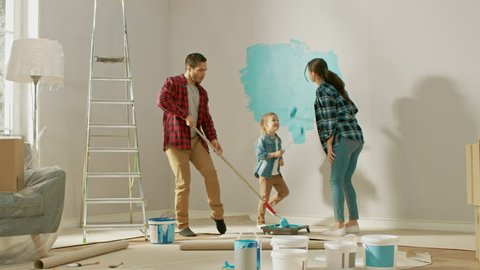 Family Time Together with Small Daughter. Young Father and Mother are Dancing and Fooling Around with Paint Rollers. Wall Paint Color is Light Blue. Room at Home is Prepared for Renovations.