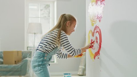 Happy Little Girl with Hands Dipped in Yellow and Red Paint Draws a Colorful Heart with Smile on the Wall. She is Having Fun and Laughs. Home is Being Renovated.