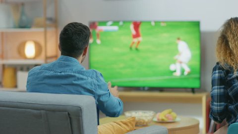 At Home Young Couple Watches Soccer Match on TV, They Worry very Emotionally, Guy Aggressively Gesticulates and They are Devasted after Their Team Misses the Goal. Back View Camera Shot.