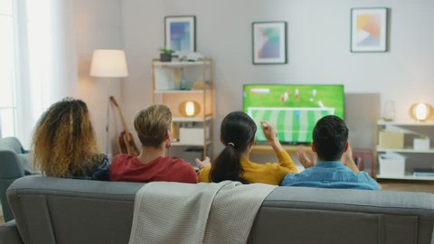 At Home Diverse Group of Sports Fans Sitting on a Couch Watching Important Soccer Match on TV, They Cheer for the Team, Celebrate Victory after Team Scoring Winning Goal. Cozy Room with Snacks, Drinks