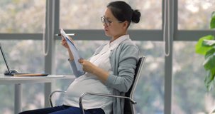 One pregnant Chinese business woman working in the office, looking on paperworks with laptop on table by the window, female business lady working late into pregnancy modern working woman 4k clip