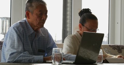 Side view of old senior Asian couple using multimedia devices in a comfortable home. They are sitting together at the dining table, Social distancing and self isolation in quarantine 