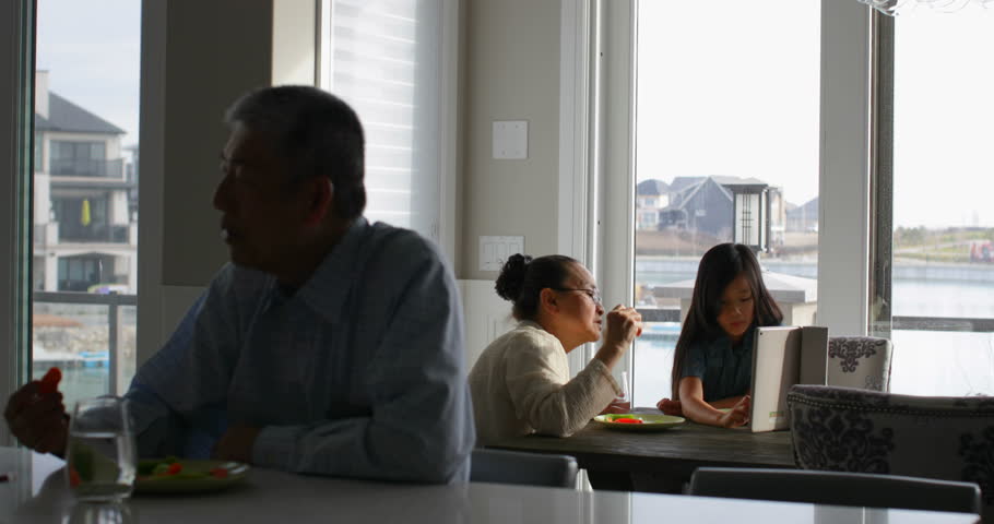 Front view of multi-generation Asian family eating food at dining table in a comfortable home. Granddaughter using digital tablet  | Shutterstock HD Video #1023204109
