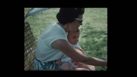 Mom feeds baby boy sitting outside on lawn chair 1960
