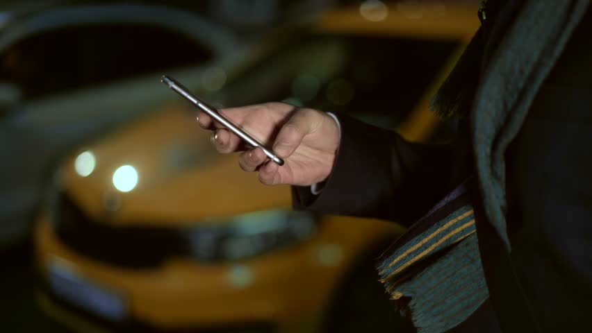 Concept of taxi calling service. Close up slow motion shot of male hands clicking on the phone, observing the map of the city. Left to right pan shot bakcground of yellow taxi at night