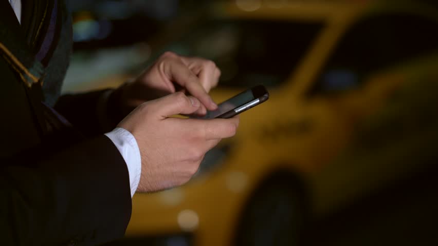Concept of taxi calling service. Close up slow motion shot of male hands clicking on the phone, observing the map of the city. Right to left pan shot bakcground of yellow taxi at night