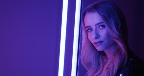 Portrait of the beautiful blonde young woman standing among neon lights and smiling cheerfully to the camera. Close up.