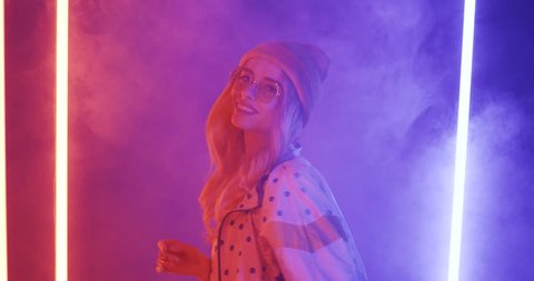 Cheerful stylish blonde girl in hat and sunglasses dancing and smiling in the smoke and neon lights.