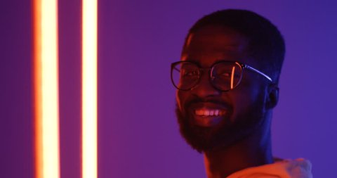 Close upof teh profile of the young African American man in glasses turning his head to the camera and smiling. Portrait. Neon lights.