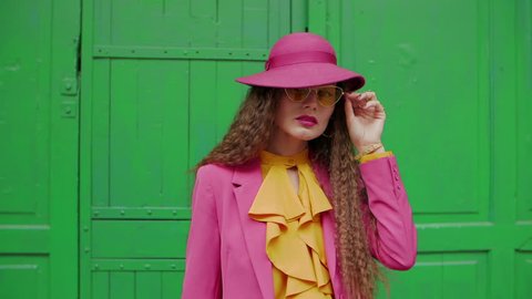 Young fashionable woman with long beautiful curly hair turns around. Model wearing trendy yellow sunglasses, pink hat, blazer, yellow blouse, hoop earrings