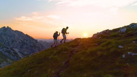 AERIAL, LENS FLARE: Active young tourists hiking up a grassy mountain in the Alps at sunset. Carefree hiker couple enjoying a relaxing evening trip in the breathtaking Julian Alps. Tourists on a trek.