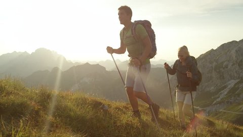 SLOW MOTION, LENS FLARE: Golden spring sun rays shine on cheerful young couple hiking up a steep grassy hill in the picturesque Alps. Smiling woman and man enjoying a hike in the stunning mountains.