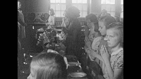1930s: Young girls stand at table with hands clasped. Line of girl walks into room to join them. Nuns usher girls into room. Girls stand praying next to set table.