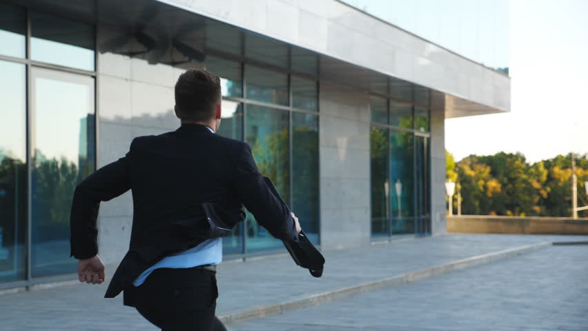 Unrecognizable businessman with briefcase runs down city street. Business man late for meeting. Successful man in suit jogging near modern building. Guy in hurry to appointment. Back view Slow motion Royalty-Free Stock Footage #1023267136