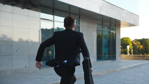 Unrecognizable businessman with briefcase runs down city street. Business man late for meeting. Successful man in suit jogging near modern building. Guy in hurry to appointment. Back view Slow motion