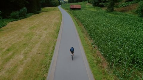 Epic aerial drone footage of professional cyclist riding down beautiful countryside road between fields and forest. Atheltic training or strength workout on sunny summer day. Healthy cycling lifestyle