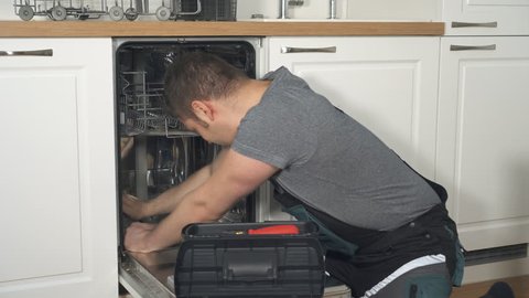 Professional handyman in overalls repairing domestic dishwasher in the kitchen.