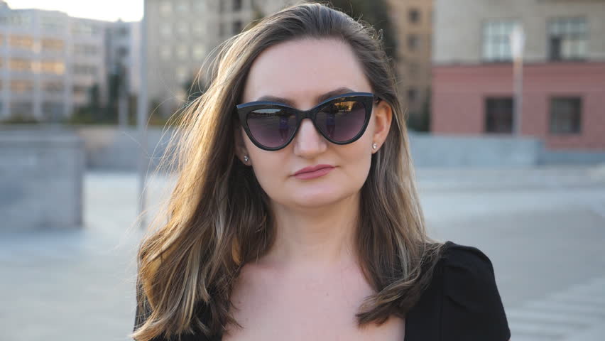 Portrait of young businesswoman in sunglasses walking in city street. Attractive business woman looking at camera. Face of confident girl commuting to work. Blurred background.Close up Slow motion