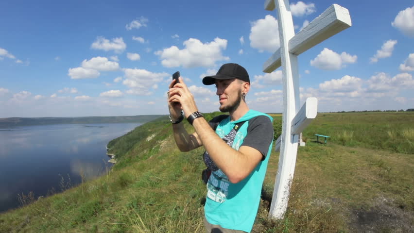 Young attractive man smiling and shooting beautiful landscapes on his smartphone. Two more men standing by and looking at a huge lake.
