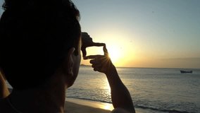 Young man with curly hair making photo frame with his hands at amazing sunset on the beach. Silhouette of photographer. Art inspiration and beautiful light.