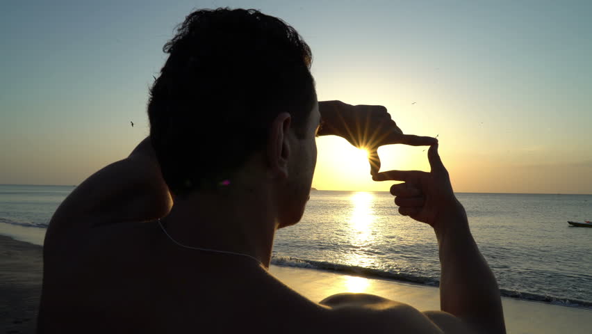 Young man with curly hair making photo frame with his hands at amazing sunset on the beach. Silhouette of photographer. Art inspiration and beautiful light.