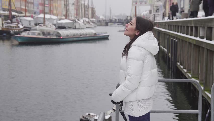 Youg beautiful woman stands at a Nyhavn pier and looks happy at winter in Copenhagen, Denmark with a pleasure boat sails by (4K, 25fps)